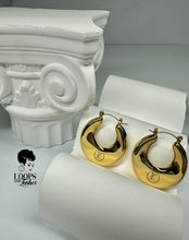 Load image into Gallery viewer, A pair of gold Dynasty Hoops with a sleek, minimalist hollow design, crafted from stainless steel with luxurious PVD gold plating. These water-resistant hoops, engraved with a signature submark logo, symbolize timeless elegance and strength. Perfect for everyday wear or statement styling, they honor the legacy of bold, stylish women across generations.