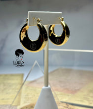 Load image into Gallery viewer, A pair of gold Dynasty Hoops with a sleek, minimalist hollow design, crafted from stainless steel with luxurious PVD gold plating. These water-resistant hoops, engraved with a signature submark logo, symbolize timeless elegance and strength. Perfect for everyday wear or statement styling, they honor the legacy of bold, stylish women across generations.