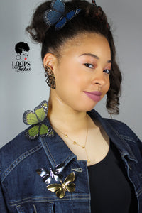 Girl wearing gold Fearless Wings butterfly earrings and a denim jacket adorned with gold and silver butterfly brooches, with a delicate butterfly resting on her shoulder. Bold jewelry styling with nature-inspired elegance