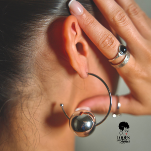 Close-up of an ear with a silver hoop earring with sphere, hand near earring, and 'Loops and Lashes' logo.
